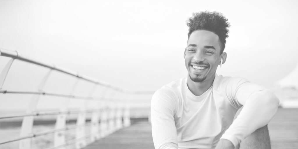 Cheerful Curly Dark Skinned Man In White Long Sleeved T Shirt Smiles Sincerely And Rests Near Sea