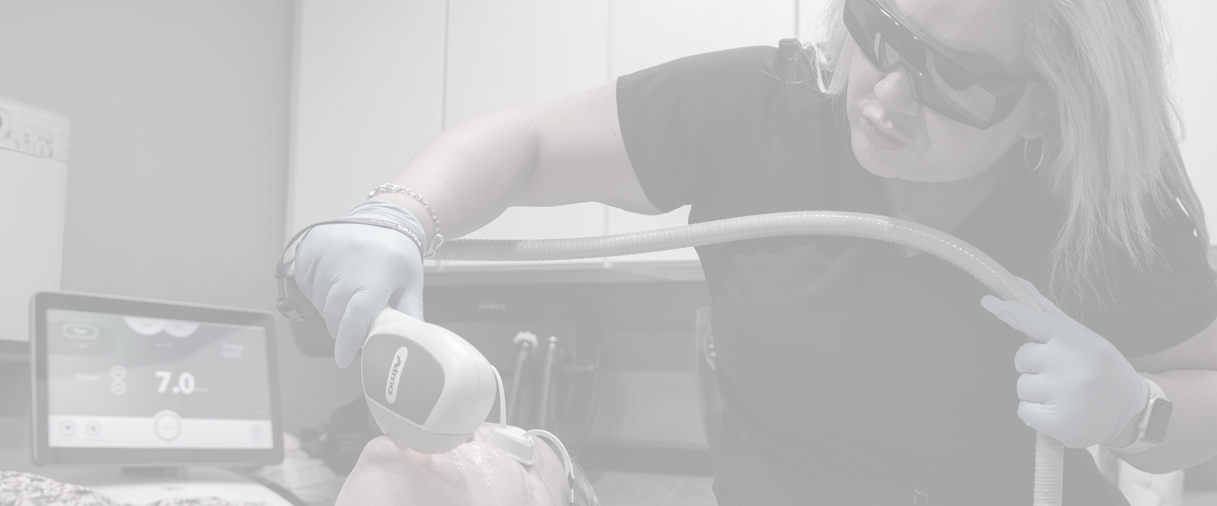 The aesthetician is holding a handheld device, which appears to be a laser or IPL (Intense Pulsed Light) machine, and applying it to the patient's face.